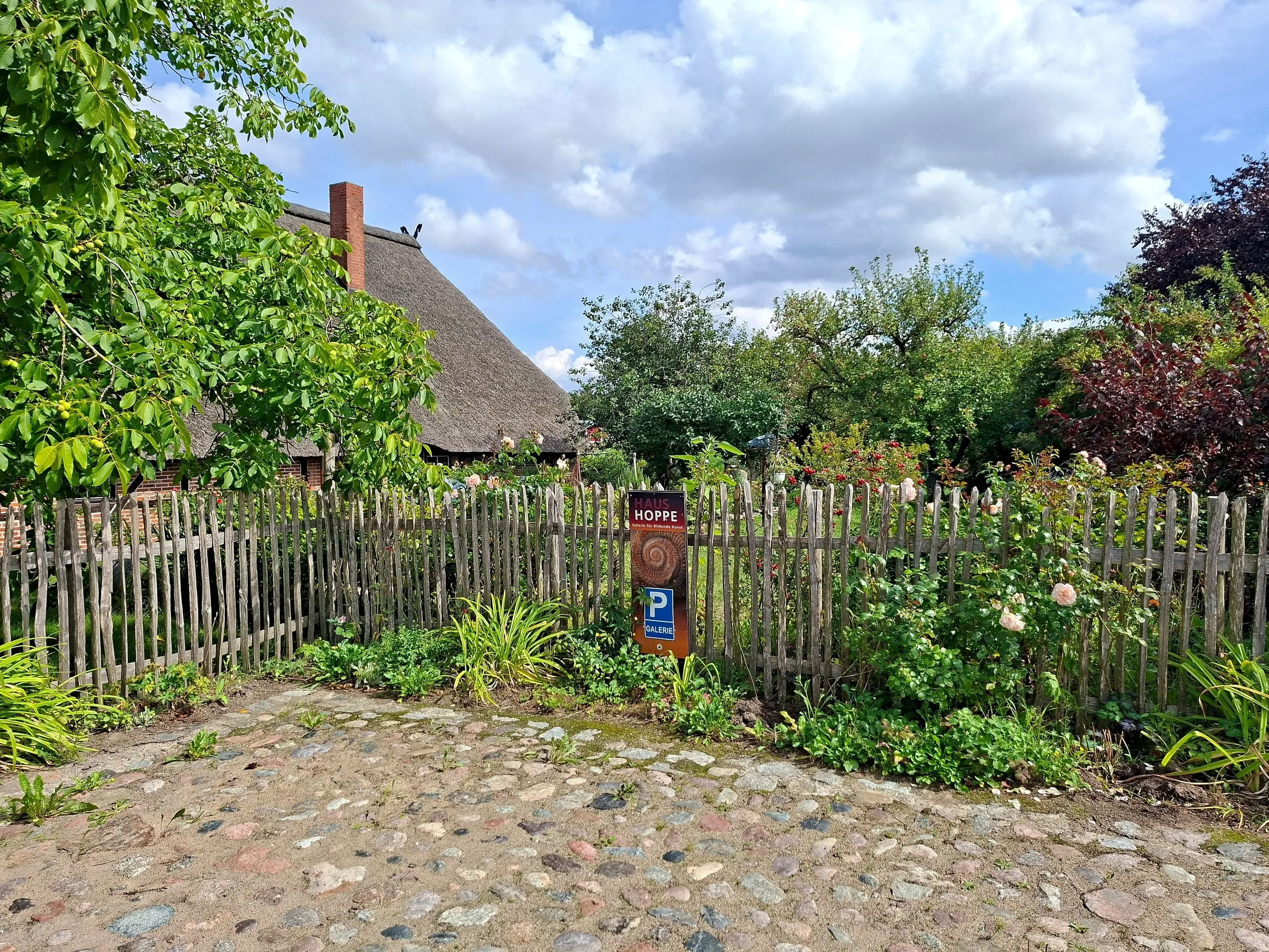 Bauerngarten hinter dem Staketenzaun, mit Kopfsteinweg und dem Schild HAUS HOPPE