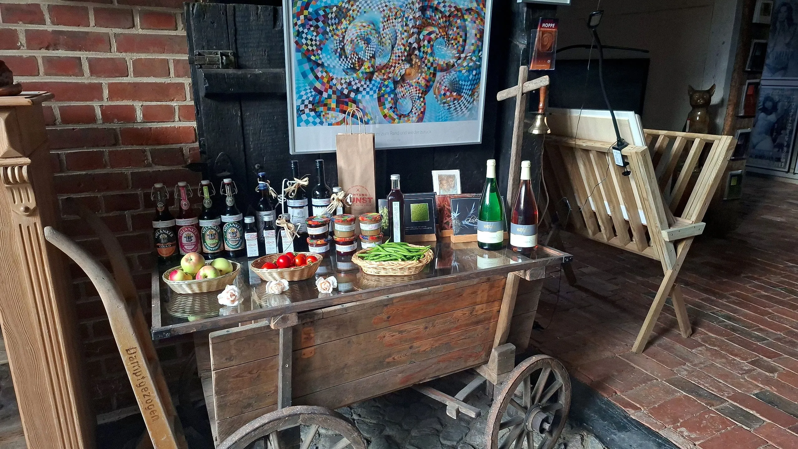 Alter Marktwagen mit Glasplatte im Hofladen, darauf Bier, Marmelade, Öle und Körbe mit Äpfeln, Tomaten und Bohnen, dahinter ein großes farbiges Kunstwerk an der Backsteinwand